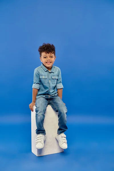 Happy and curly african american preschooler boy in denim clothes sitting on white cube on blue — Stock Photo