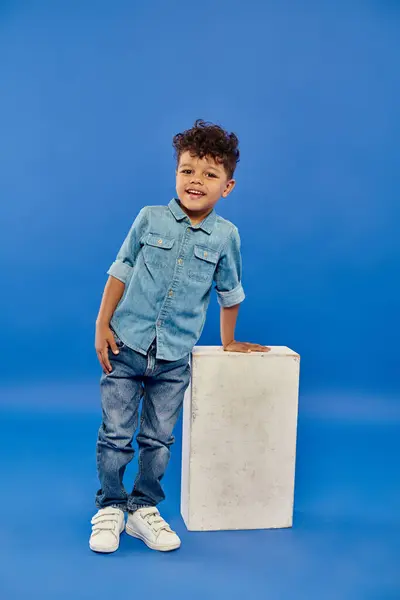 Happy and curly african american preschooler boy in denim clothes leaning on white cube on blue — Stock Photo