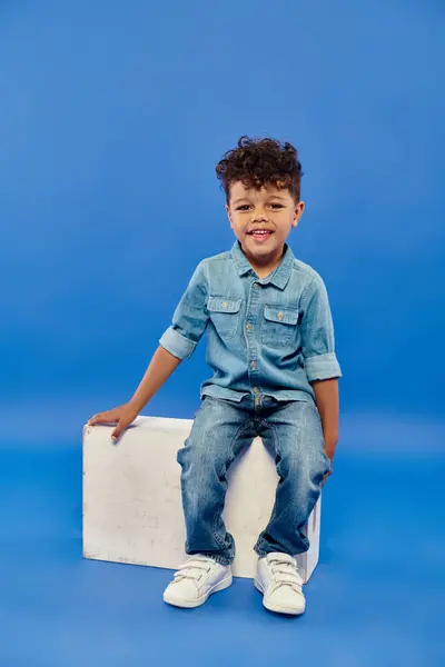 Веселый и кудрявый африканский дошкольник в джинсовой одежде, сидящий на белом кубе на голубом — стоковое фото
