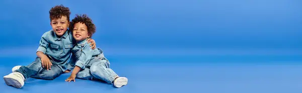 Adorável afro-americanos irmãos em roupas jeans elegantes sentado e abraçando em azul, bandeira — Fotografia de Stock