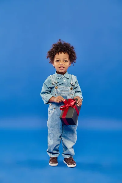 Happy african american boy in denim clothes standing with Christmas present on blue backdrop — Stock Photo