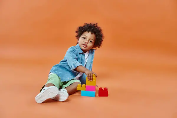 Carino ragazzo bambino africano americano in camicia di jeans seduto e giocare blocchi di costruzione su arancione — Foto stock