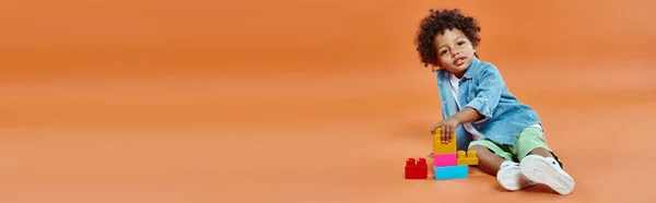 African american toddler boy in denim shirt sitting and playing building blocks on orange, banner — Stock Photo