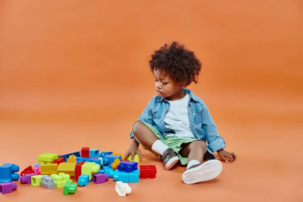 Adorabile ragazzo afroamericano bambino in camicia di jeans seduto e giocare blocchi di costruzione su arancione — Foto stock