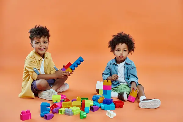 Happy african american siblings boy in casual attire sitting and playing building blocks on orange — Stock Photo