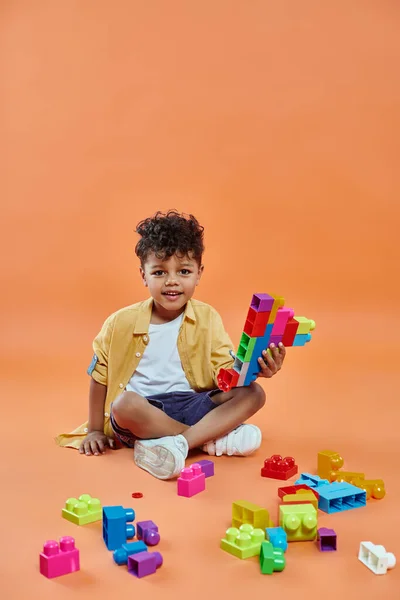 Fröhlicher afrikanisch-amerikanischer Junge in lässiger Kleidung sitzt und spielt bunte Bausteine auf orange — Stockfoto