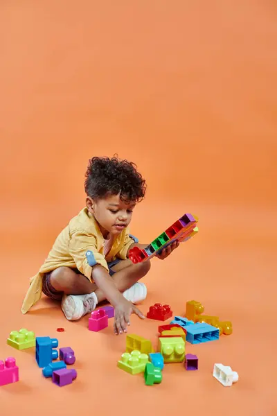 Felice ragazzo africano americano in abbigliamento casual seduto e giocando mattoni su sfondo arancione — Foto stock
