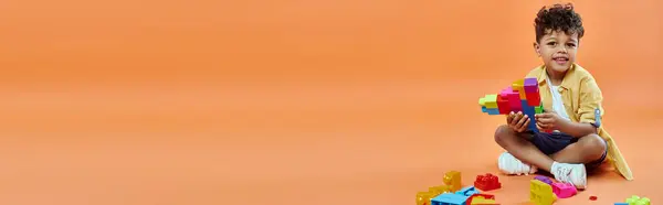 Счастливый африканский американец в повседневной одежде сидит и играет строительные блоки на оранжевый, баннер — стоковое фото