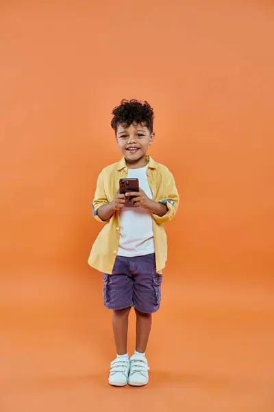 Heureux garçon préscolaire afro-américain en tenue décontractée en utilisant smartphone sur fond orange — Photo de stock