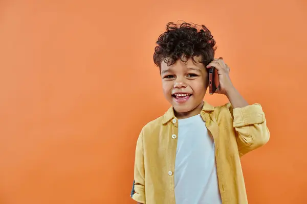 Glücklich afrikanisch-amerikanischer Vorschuljunge in lässiger Kleidung, der auf einem Smartphone auf orangefarbenem Hintergrund spricht — Stockfoto