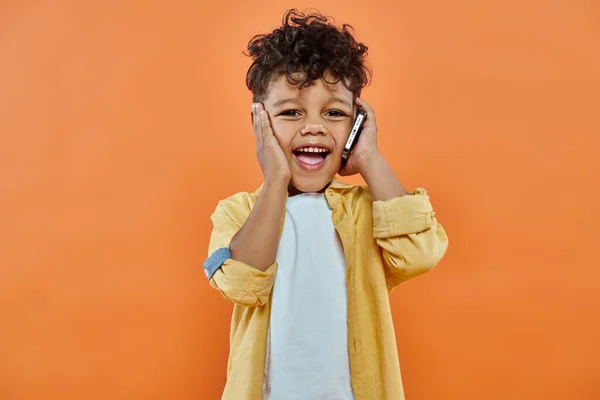 Joyful african american preschooler boy in casual attire talking on smartphone on orange background — Stock Photo