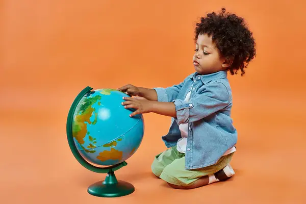 Happy african american toddler boy in casual attire touching globe and sitting on orange backdrop — Stock Photo
