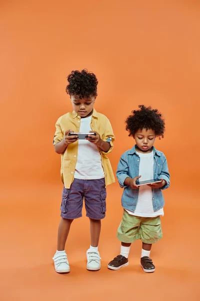 Smiling african american siblings in casual summer attire playing on smartphones on orange — Stock Photo
