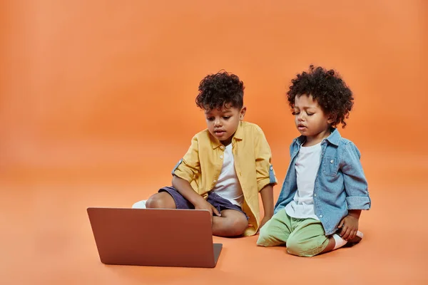 Adorable african american siblings in casual summer attire watching movie on laptop, orange backdrop — Stock Photo