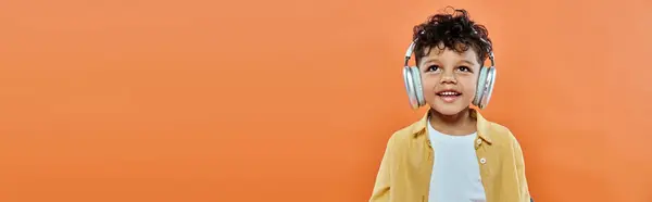 Ragazzo afroamericano felice e riccio che ascolta musica in cuffie senza fili su arancione, banner — Foto stock