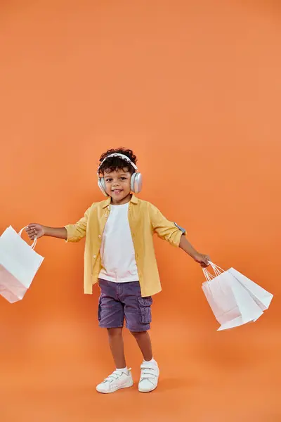 Gioioso ragazzo afroamericano che ascolta musica in cuffia e tiene borse della spesa su arancione — Foto stock