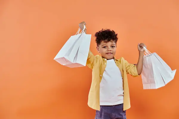 Happy african american boy in casual attire smiling and holding shopping bags on orange background — Stock Photo