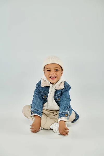 Happy african american preschooler boy in winter attire and beanie hat sitting on grey background — Stock Photo