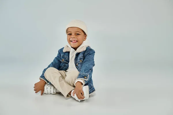 Радісний афроамериканський хлопчик-дошкільник в зимовому вбранні і капелюсі бані сидить на сірому фоні — стокове фото