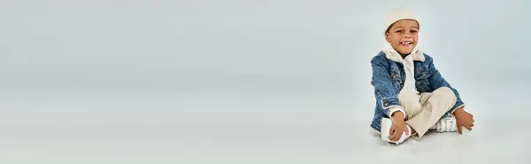 Felice ragazzo prescolastico africano americano in abbigliamento invernale e cappello beanie seduto sul grigio, banner — Foto stock
