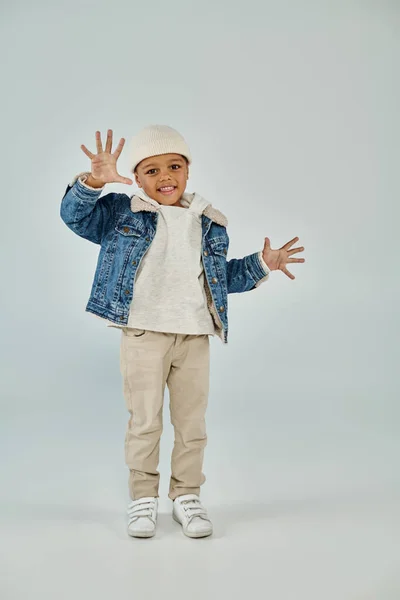 Cheerful african american preschooler boy in winter attire and beanie hat gesturing on grey backdrop — Stock Photo