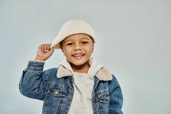 Happy african american preschooler boy in winter attire pulling beanie hat on grey backdrop — Stock Photo