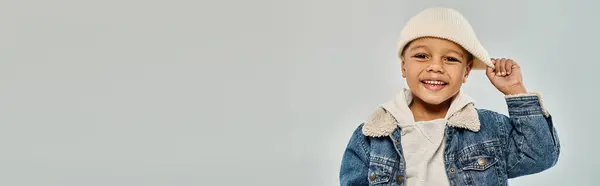 Felice ragazzo prescolastico africano americano in abbigliamento invernale che tira cappello beanie su sfondo grigio, banner — Foto stock