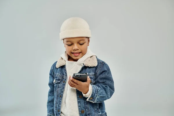African american preschooler boy in winter attire and beanie hat using smartphone on grey backdrop — Stock Photo