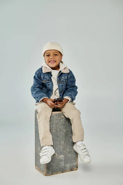 Happy african american boy in winter attire and beanie using smartphone and sitting on concrete cube — Stock Photo