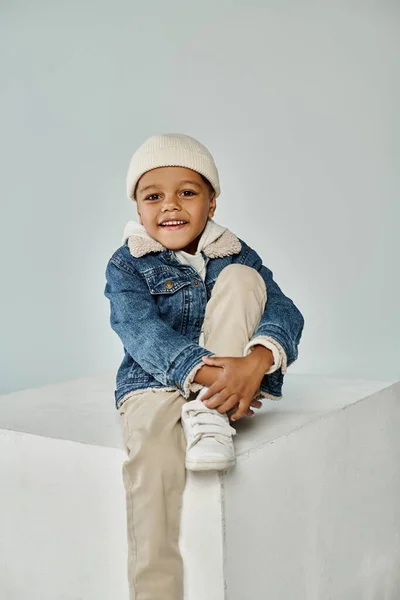 Happy african american boy in winter attire and beanie hat sitting on concrete cube on grey — Stock Photo