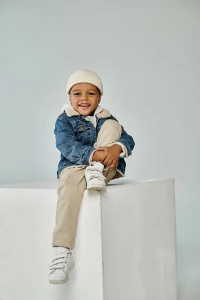 Cute happy african american boy in winter attire and beanie hat sitting on concrete cube on grey — Stock Photo