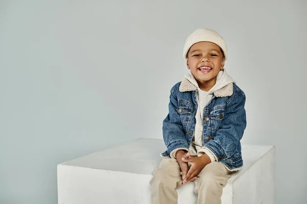 Carino gioioso ragazzo africano americano in abbigliamento invernale e cappello berretto seduto su cubo di cemento su grigio — Foto stock