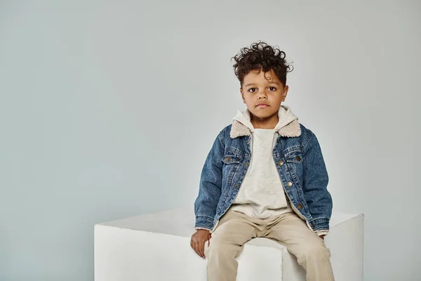 Curly african american boy in winter attire and beanie sitting on concrete cube on grey backdrop — Stock Photo