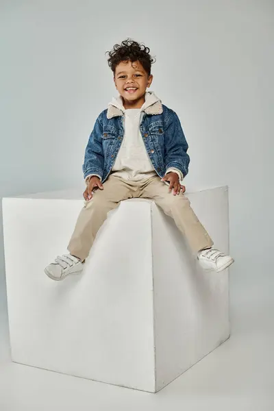 Happy african american boy in winter attire and beanie sitting on concrete cube on grey backdrop — Stock Photo