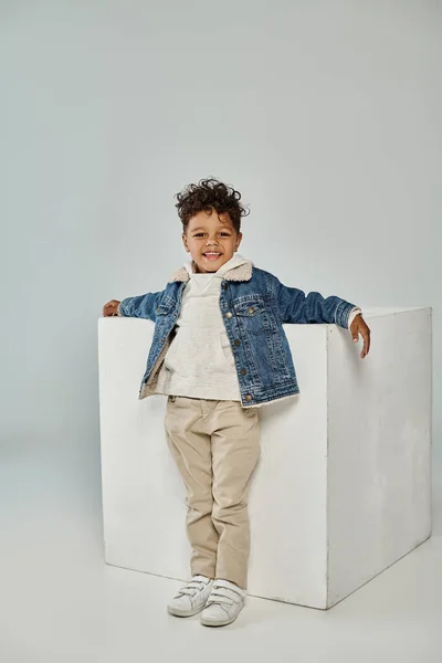 Cheerful african american boy in winter attire and beanie sitting on concrete cube on grey backdrop — Stock Photo