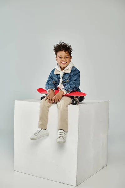 Joyeux garçon afro-américain en tenue d'hiver assis sur cube avec planche de penny sur fond gris — Photo de stock