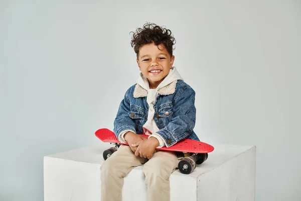 Happy african american boy in winter attire sitting on cube with penny board on grey background — Stock Photo