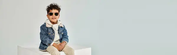Ricci ragazzo africano americano in abbigliamento invernale e occhiali da sole seduti sul cubo su sfondo grigio, banner — Foto stock
