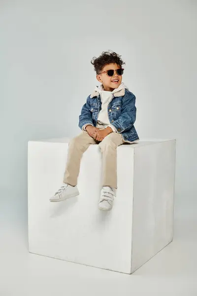 Joyeux bouclé afro-américain garçon en tenue d'hiver et lunettes de soleil assis sur cube sur fond gris — Photo de stock