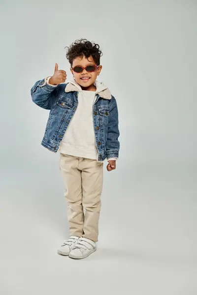 Happy curly african american boy in winter attire and sunglasses showing thumb up on grey backdrop — Stock Photo