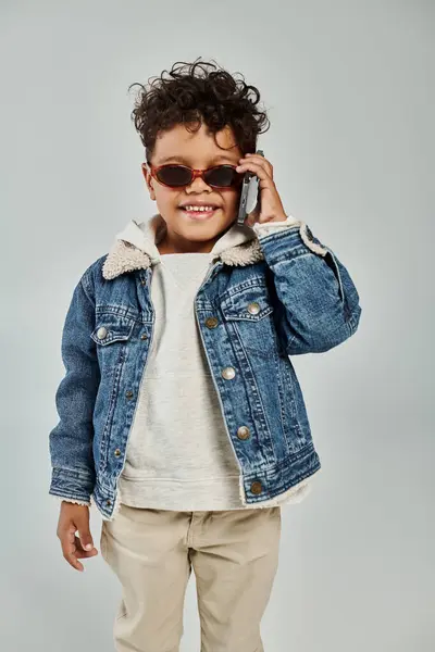 Happy african american boy in winter attire and sunglasses talking on smartphone on grey backdrop — Stock Photo