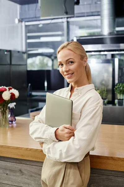 Jolie administrateur de café blonde avec ordinateur portable souriant à la caméra près du comptoir, petite entreprise — Photo de stock