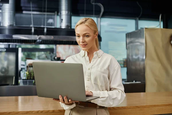 Mujer rubia sonriente mirando el ordenador portátil cerca del mostrador en la cafetería moderna, empresario en el trabajo - foto de stock