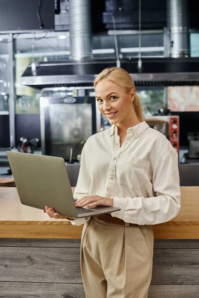 Joyeux attrayant administrateur blonde tenant ordinateur portable et regardant la caméra dans un café moderne — Photo de stock
