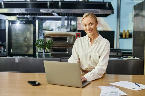Sonriente mujer rubia que trabaja en el ordenador portátil cerca de teléfono inteligente en el mostrador en la cafetería moderna, pequeña empresa - foto de stock