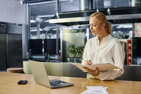 Attentive blonde woman writing in notebook near laptop and smartphone on counter in modern cafe — Stock Photo