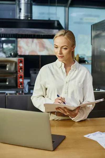 Administrateur de café souriant écrivant dans un carnet et regardant ordinateur portable sur le comptoir, petite entreprise — Photo de stock