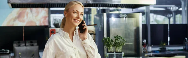 Счастливая и приветливая блондинка разговаривающая по мобильному телефону в современном кафе, горизонтальный баннер — стоковое фото