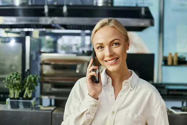 Smiling and stylish blonde woman talking on mobile phone and looking at camera in modern cafe — Stock Photo