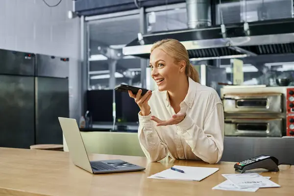 Blonde manager sending voice message on smartphone near laptop and payment terminal on cafe counter — Stock Photo